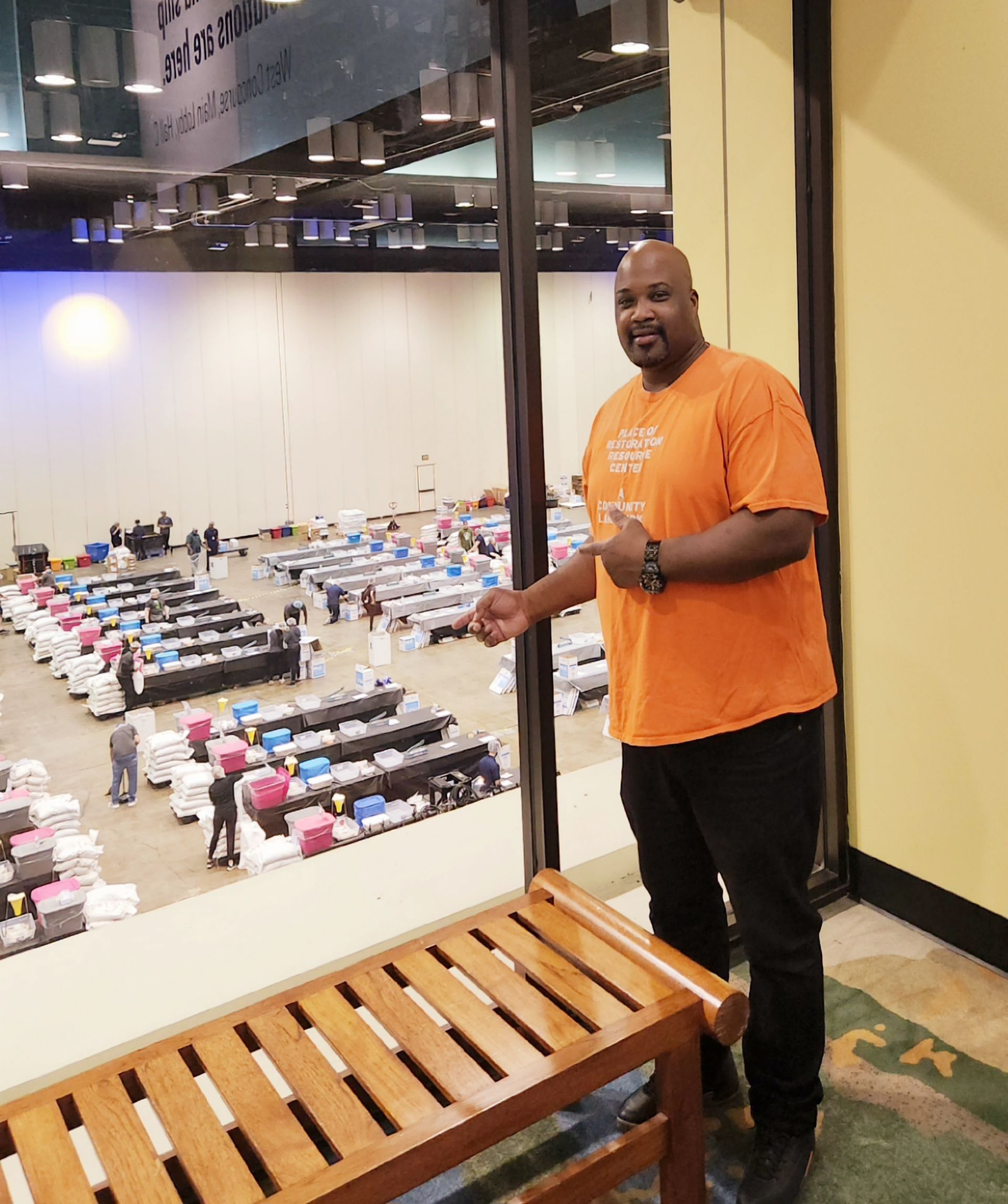 Man in orange shirt points at a large room with rows of tables, from behind a window.