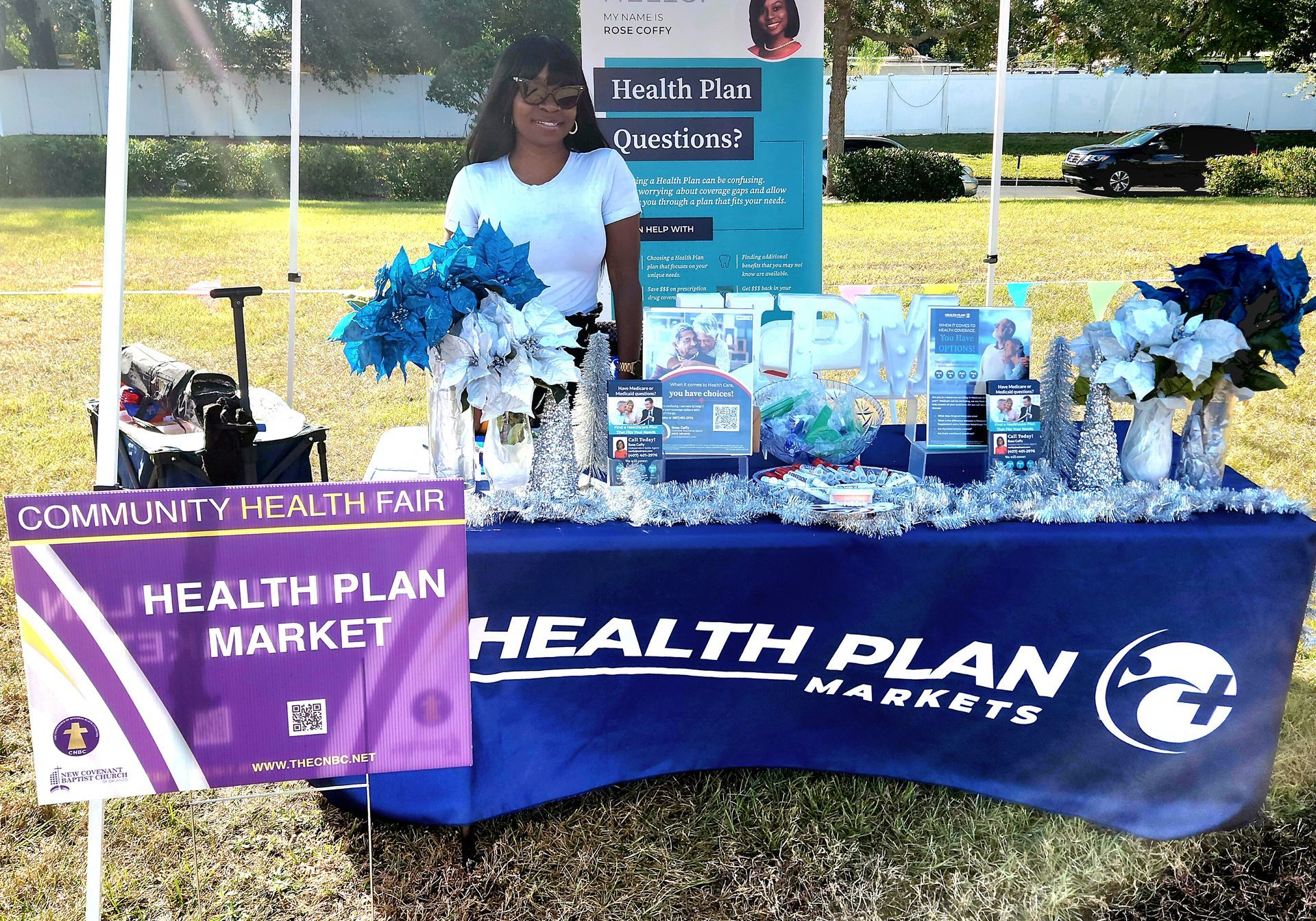 Woman at Health Plan Market booth at community health fair. Blue table, flowers, marketing materials.