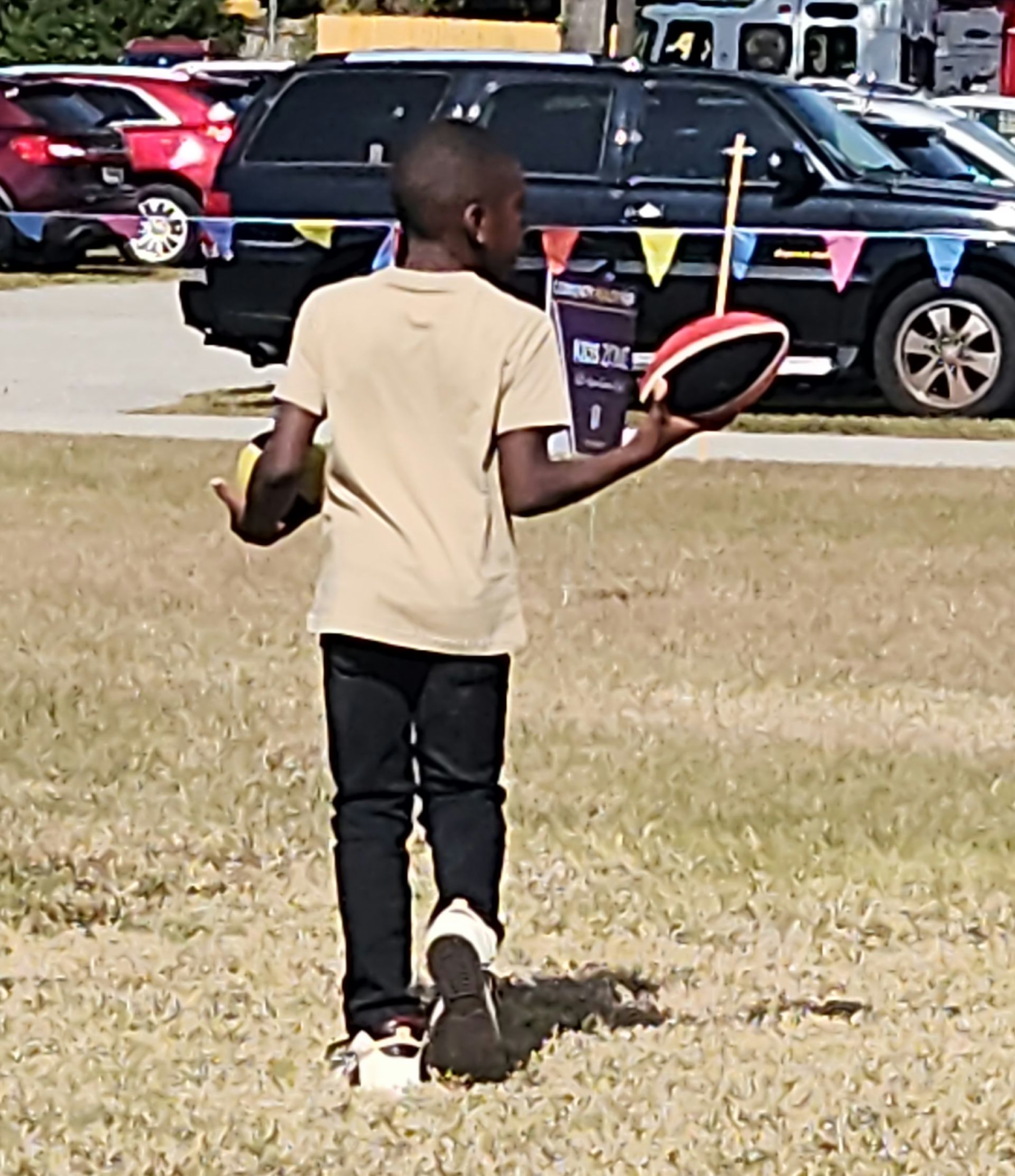 Boy walking on grass, holding a yellow ball and a football. Cars and event banners in the background.
