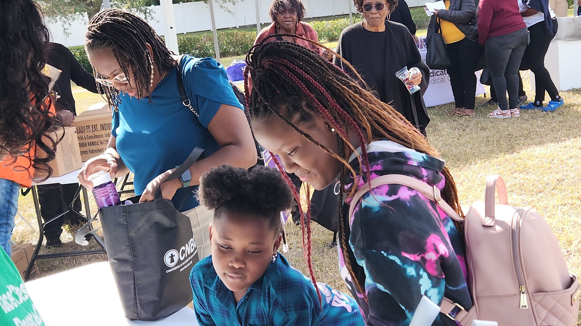 People gathered outdoors at a table, looking at items. One girl with a backpack and braided hair, others looking at handouts.