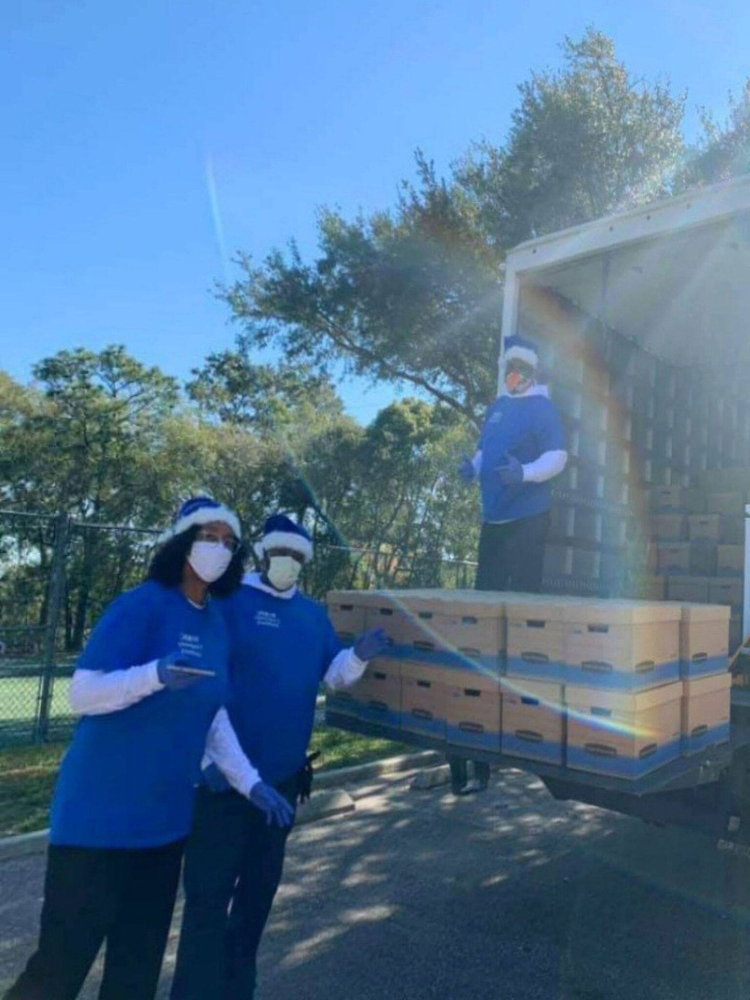 Volunteers unload boxes from a truck on a sunny day. They wear blue shirts, Santa hats, and gloves.