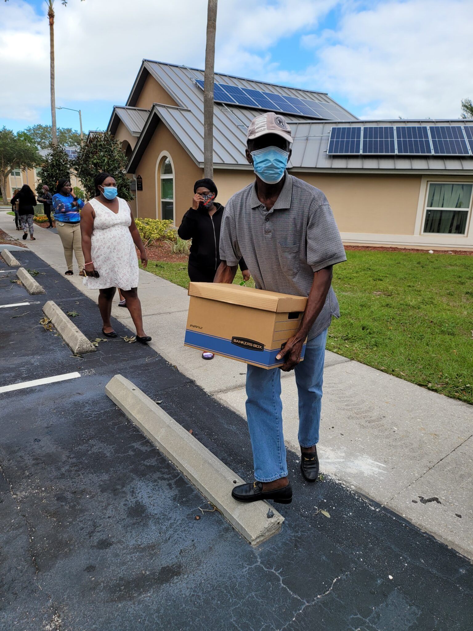 Man carrying a cardboard box in a parking lot, other people wait behind him. Buildings with solar panels in background.