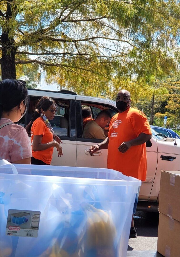 People in orange shirts unload supplies from a vehicle outside. Trees are in the background.