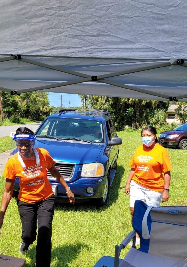 Two people in orange shirts under a tent, preparing to help a blue SUV. One wears a face shield, the other a mask.