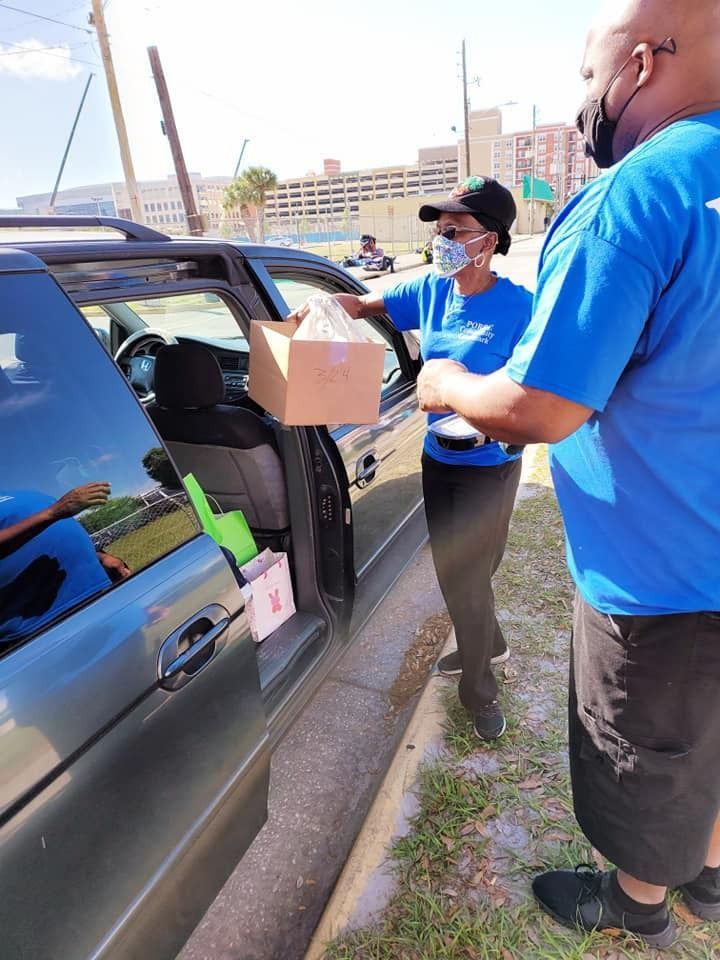 People handing food boxes from a van. They wear masks and blue shirts outdoors.