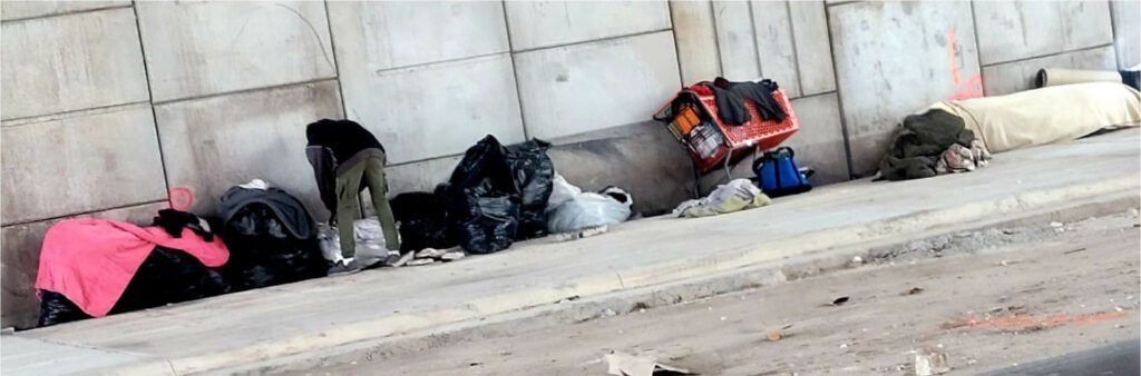 Bags and belongings are placed alongside a concrete wall. A person is seen near some of the bags.
