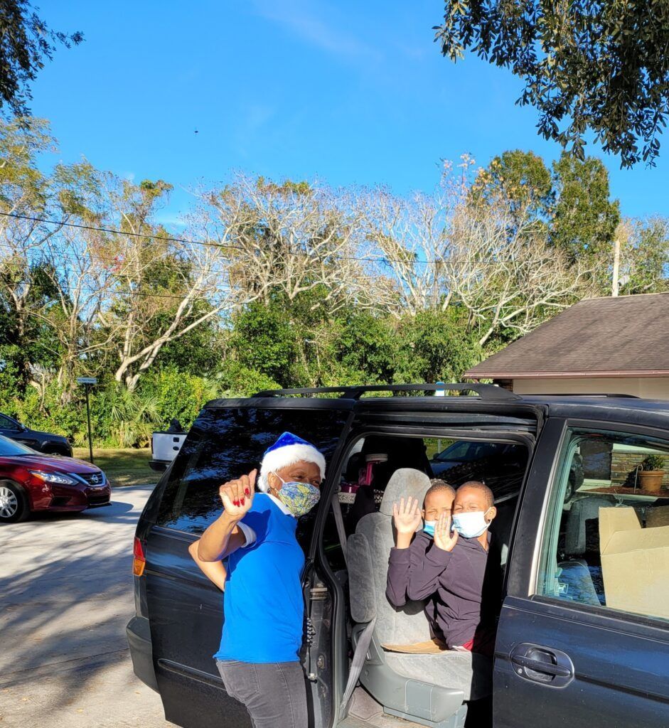 People wearing masks wave from a van. Outdoors, sunny day.