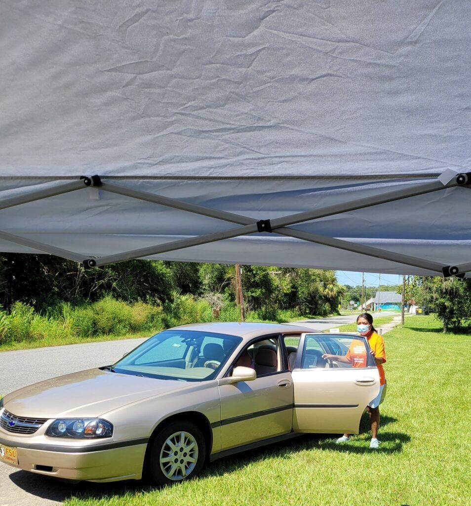A person in a mask exits a gold car under a canopy on a sunny roadside.