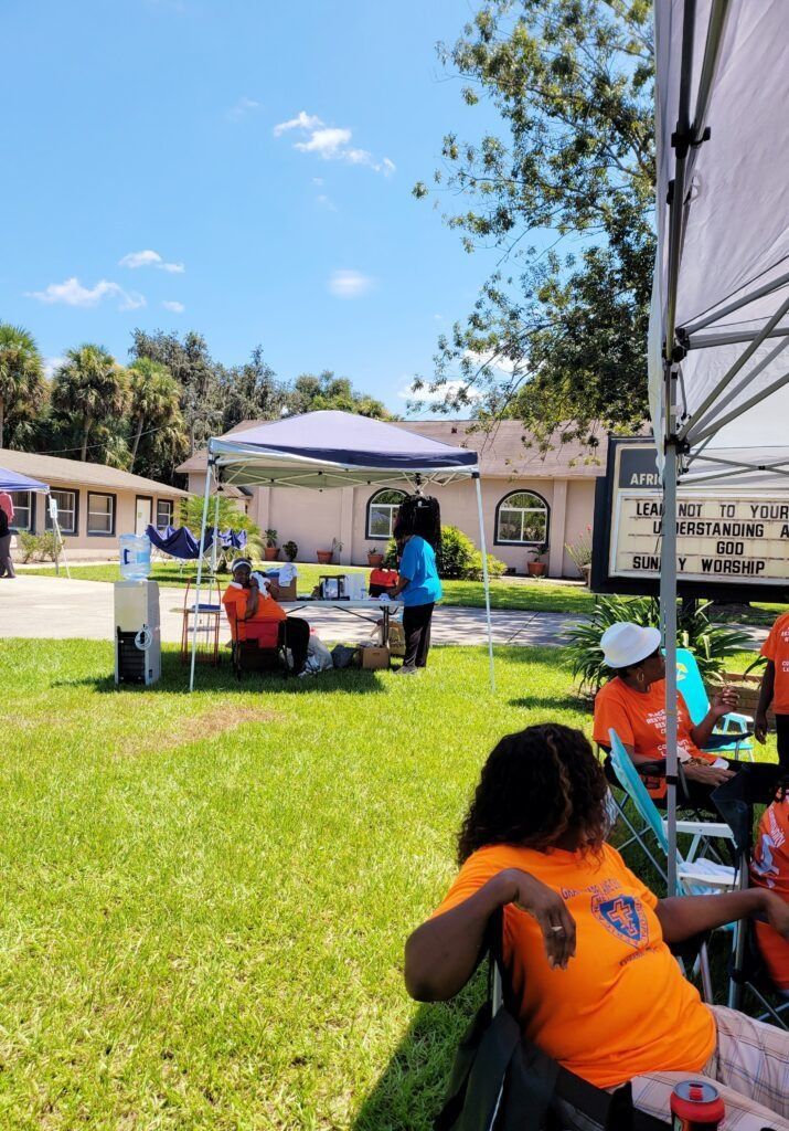 Outdoor event with people under tents, orange shirts, tables, and a building in the background.
