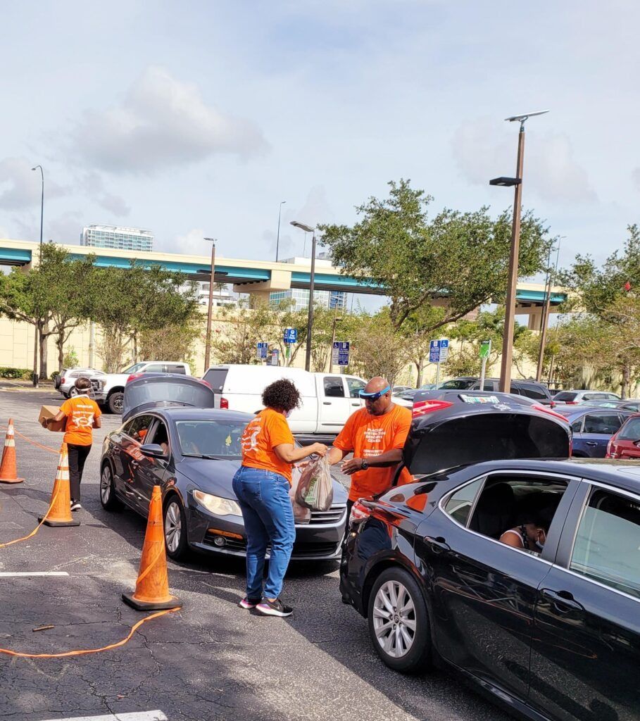Volunteers in orange shirts hand out supplies to drivers at a drive-thru event in a parking lot.