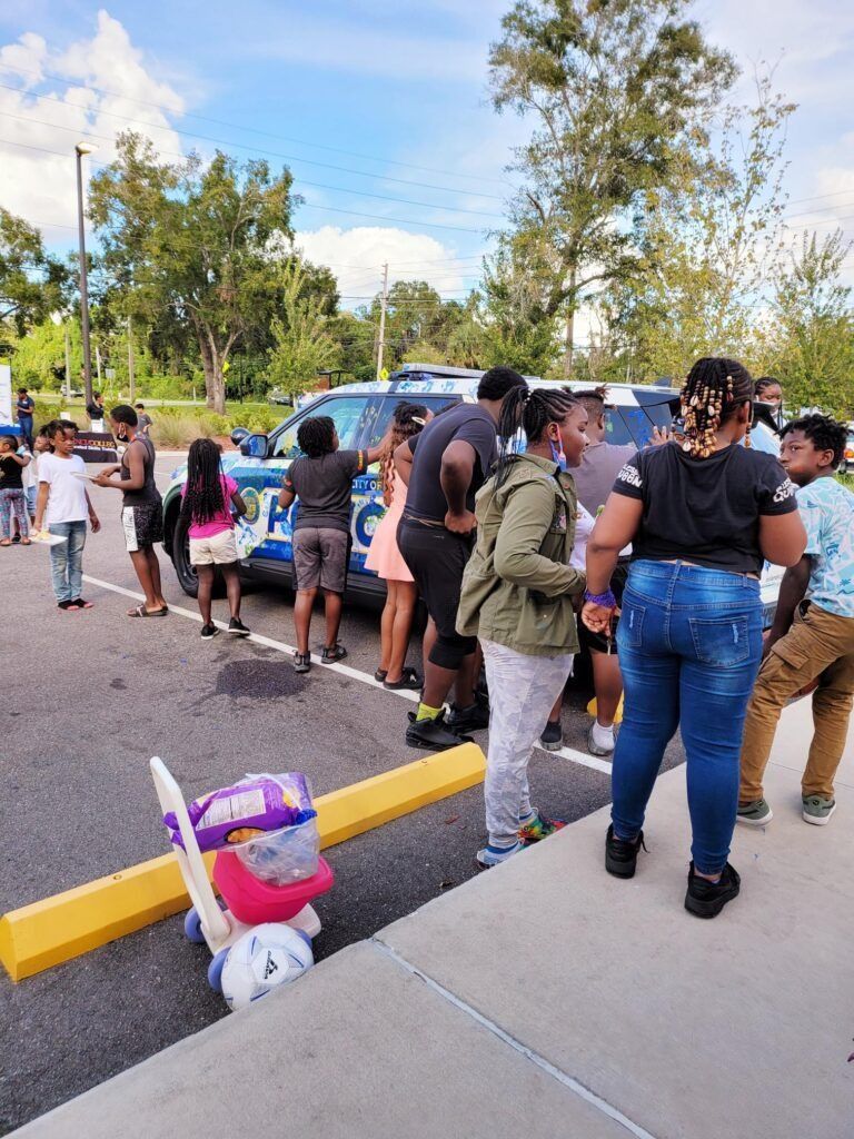 People line up near a police car. A stroller and some other items are in the foreground.