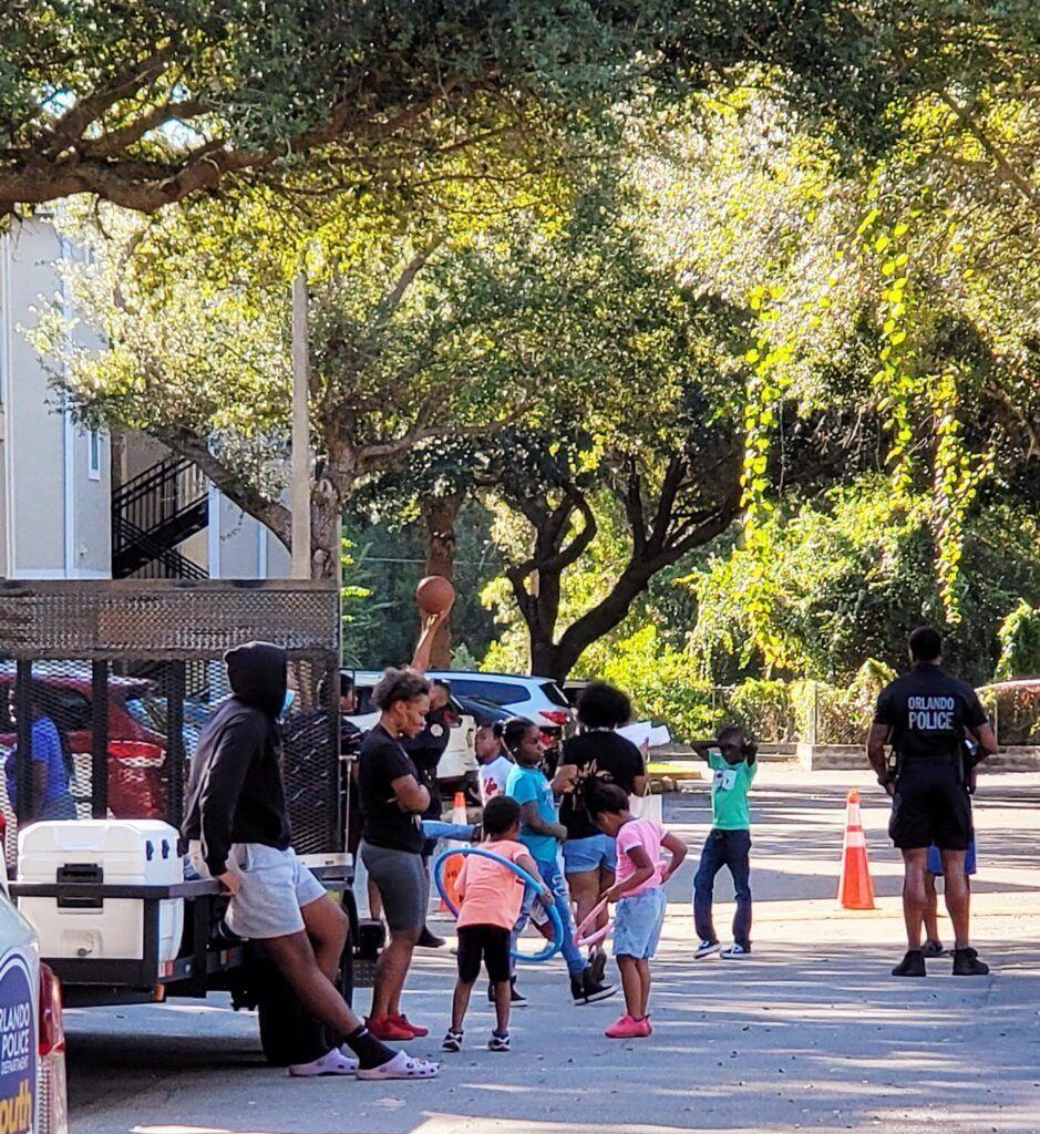 Children and adults gather outside; a police officer stands nearby. A vehicle and trees are in the background.