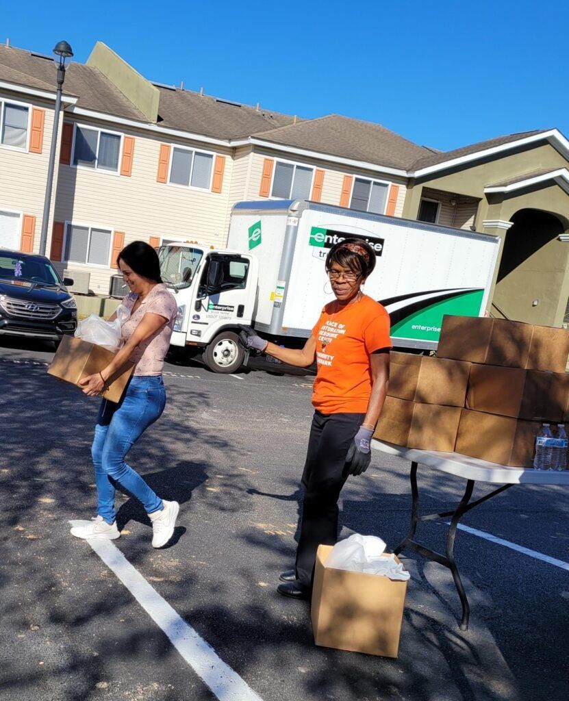 Two people unloading boxes from a truck in a parking lot. One person carries a box; another points.