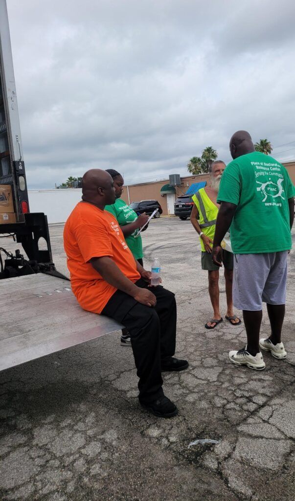 People in a parking lot, some wearing green or orange shirts, near a truck. One person is sitting. Overcast sky.