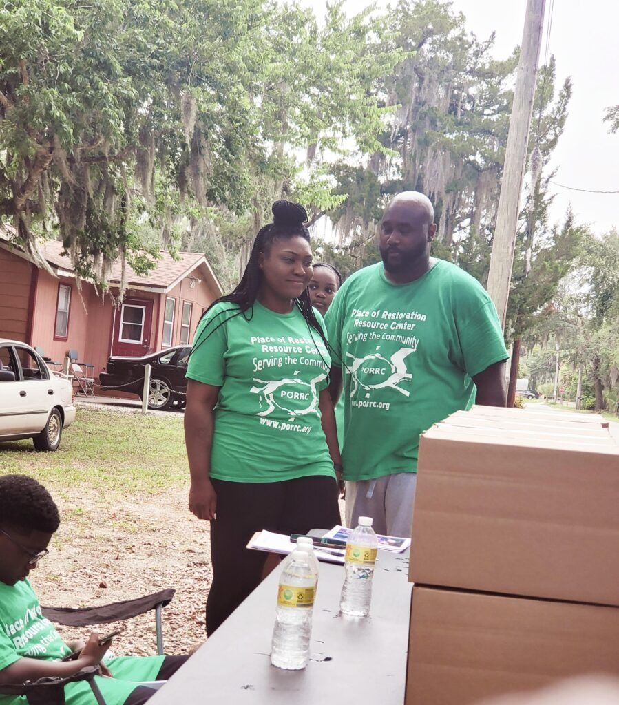 Two people in green shirts stand by a table outdoors. People in the background.