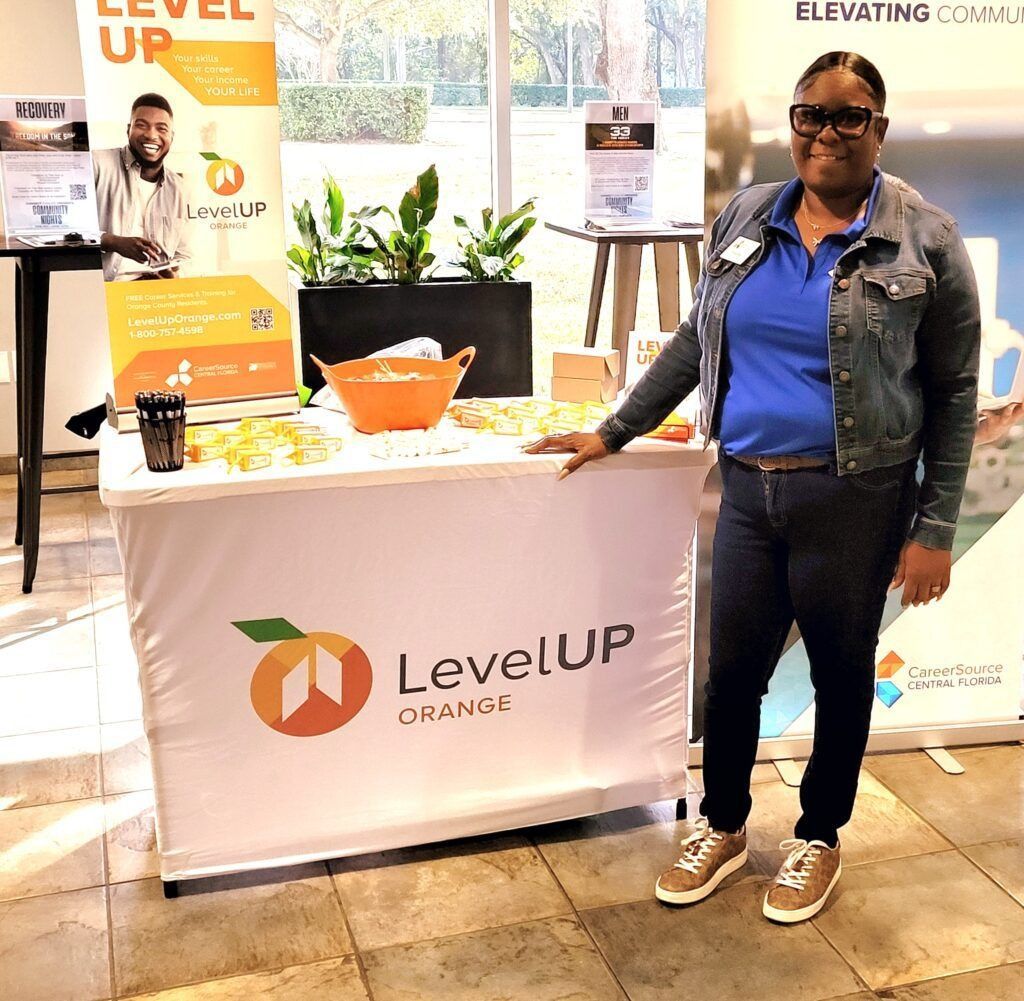 Woman stands at a "LevelUp Orange" branded table with food and plants. A banner and small sign are visible.