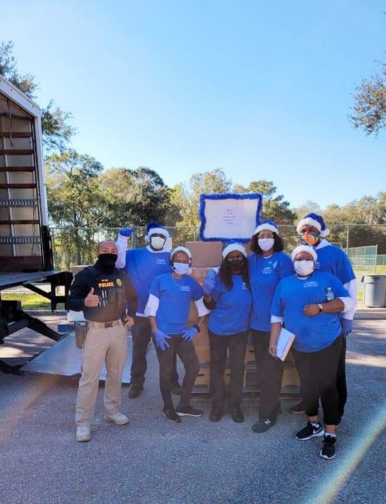 Group of people in blue shirts and Santa hats with boxes, outside. Man in uniform gives a thumbs up.
