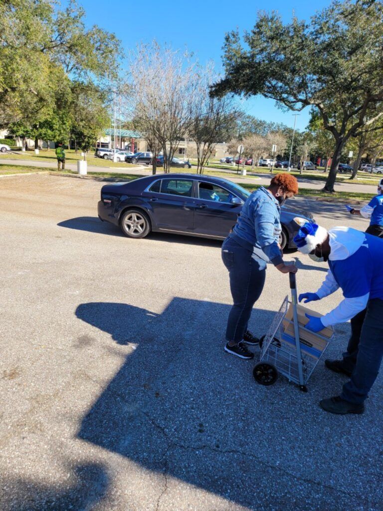 People loading groceries into a car from a cart, outdoors. Blue shirts and hats are visible.