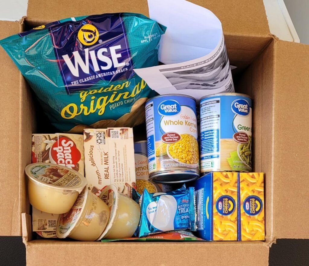 Cardboard box filled with Wise chips, canned corn and vegetables, snacks, and pudding cups.
