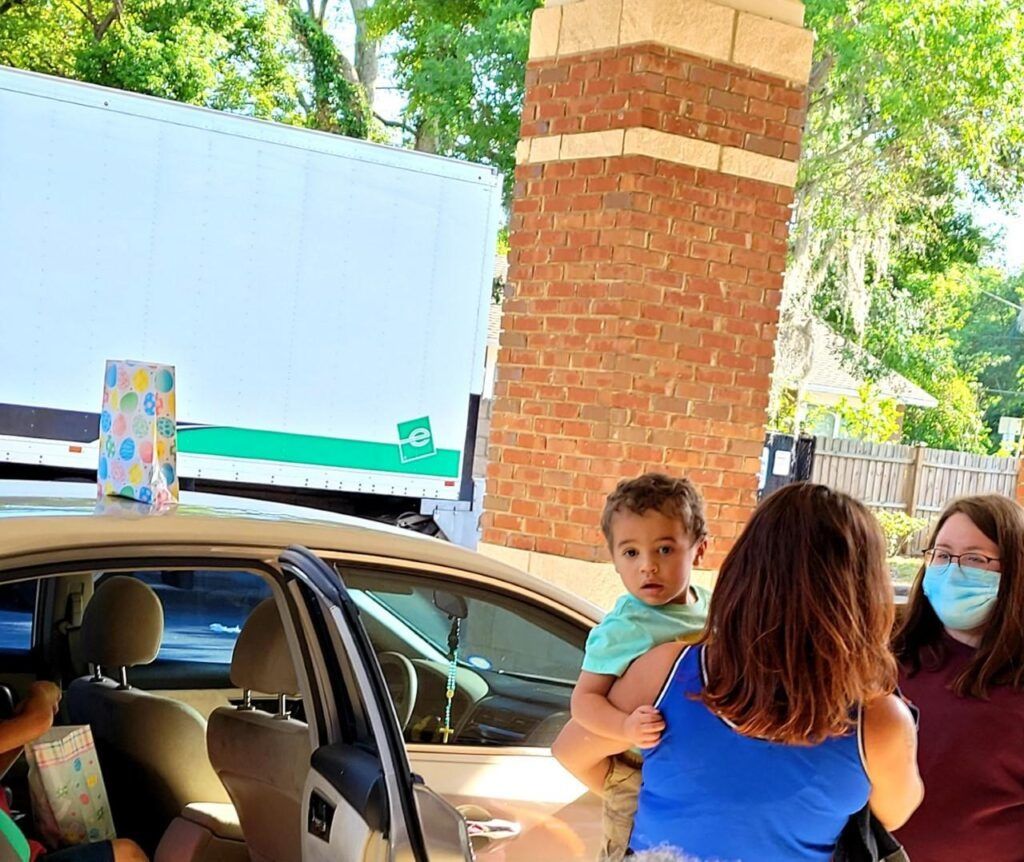 Person holding a toddler near a car, another person in a mask, a brick structure, and a truck.