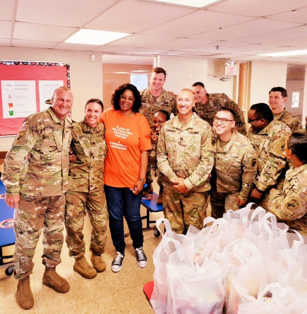 Group of soldiers and a woman in orange shirt pose with bags in a room.