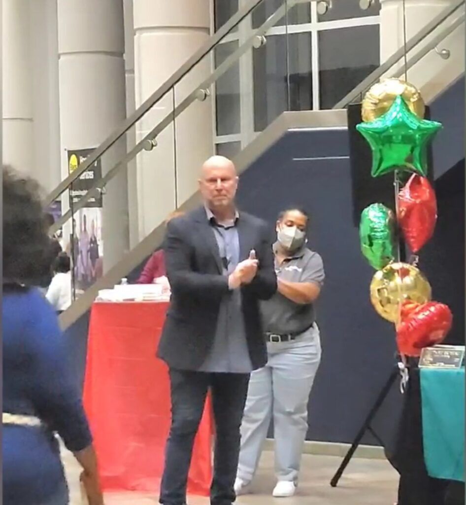 Man in a blazer applauds near balloons and a woman wearing a mask, near stairs.
