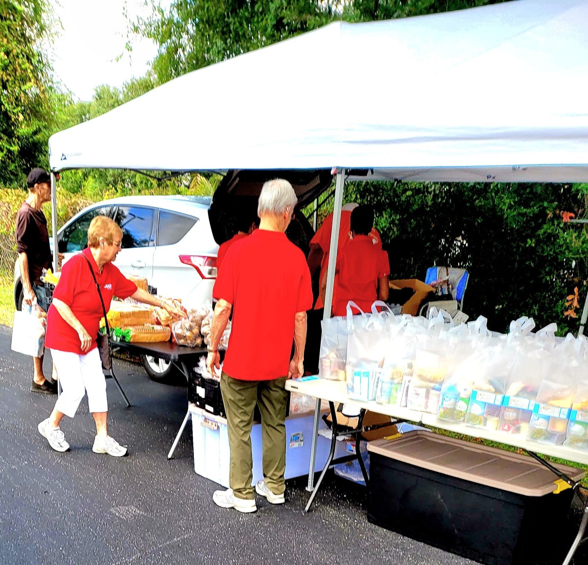 People at a roadside food stand under a white canopy. Various items are on tables; two are in red shirts.