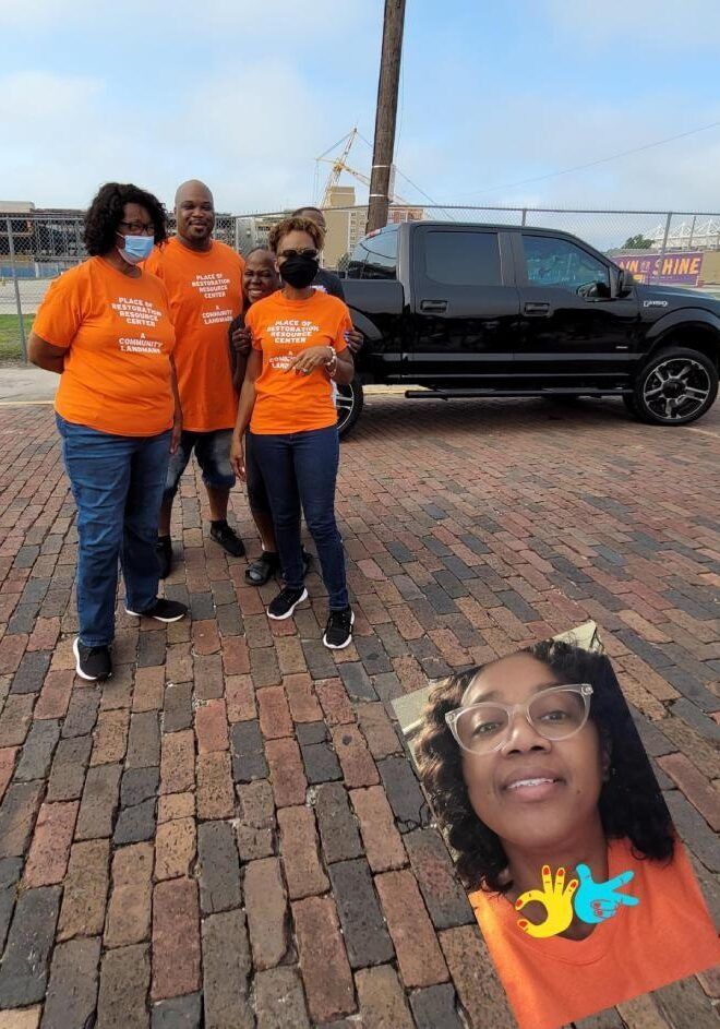 Four people in orange shirts stand near a black truck on a brick street.