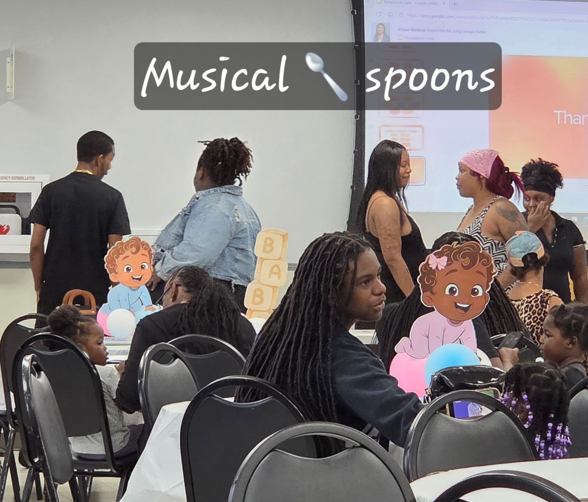 People at a lively indoor event with “Musical spoons” sign and cardboard cutout faces in the foreground.