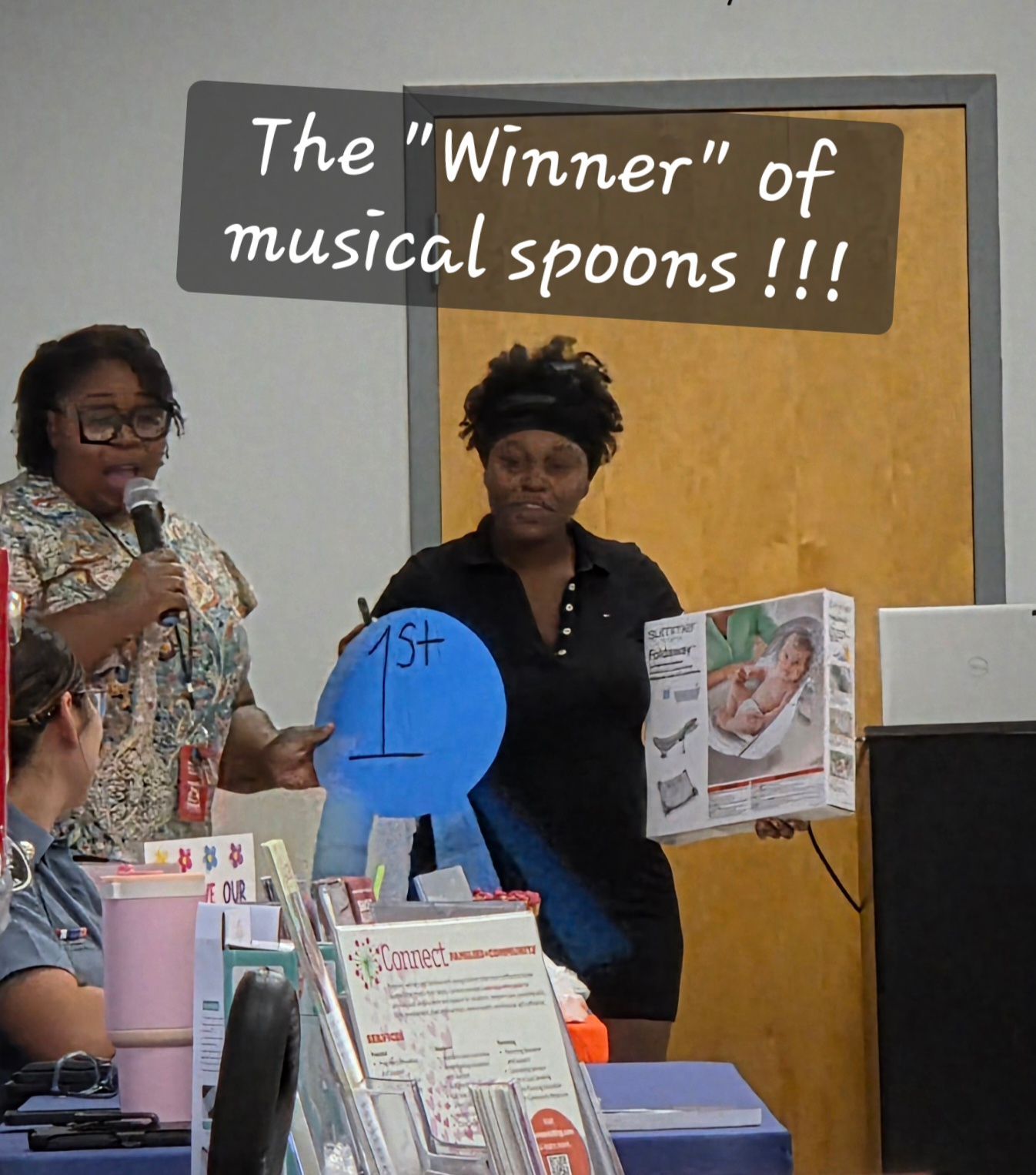 Two women beside a podium under a sign reading “The ‘Winner’ of musical spoons!!!”