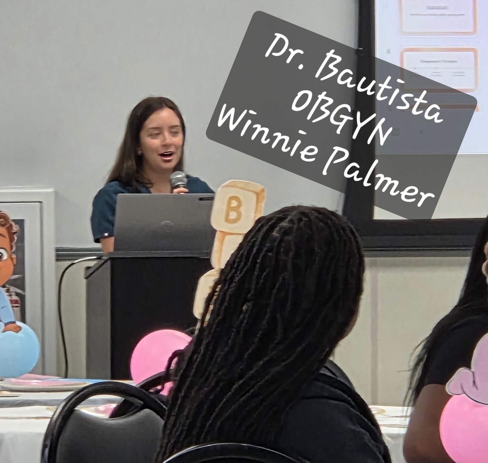 Speaker at podium in classroom, with students seated in front and a sign reading “Dr. Bautista OB-GYN Winnie Palmer”