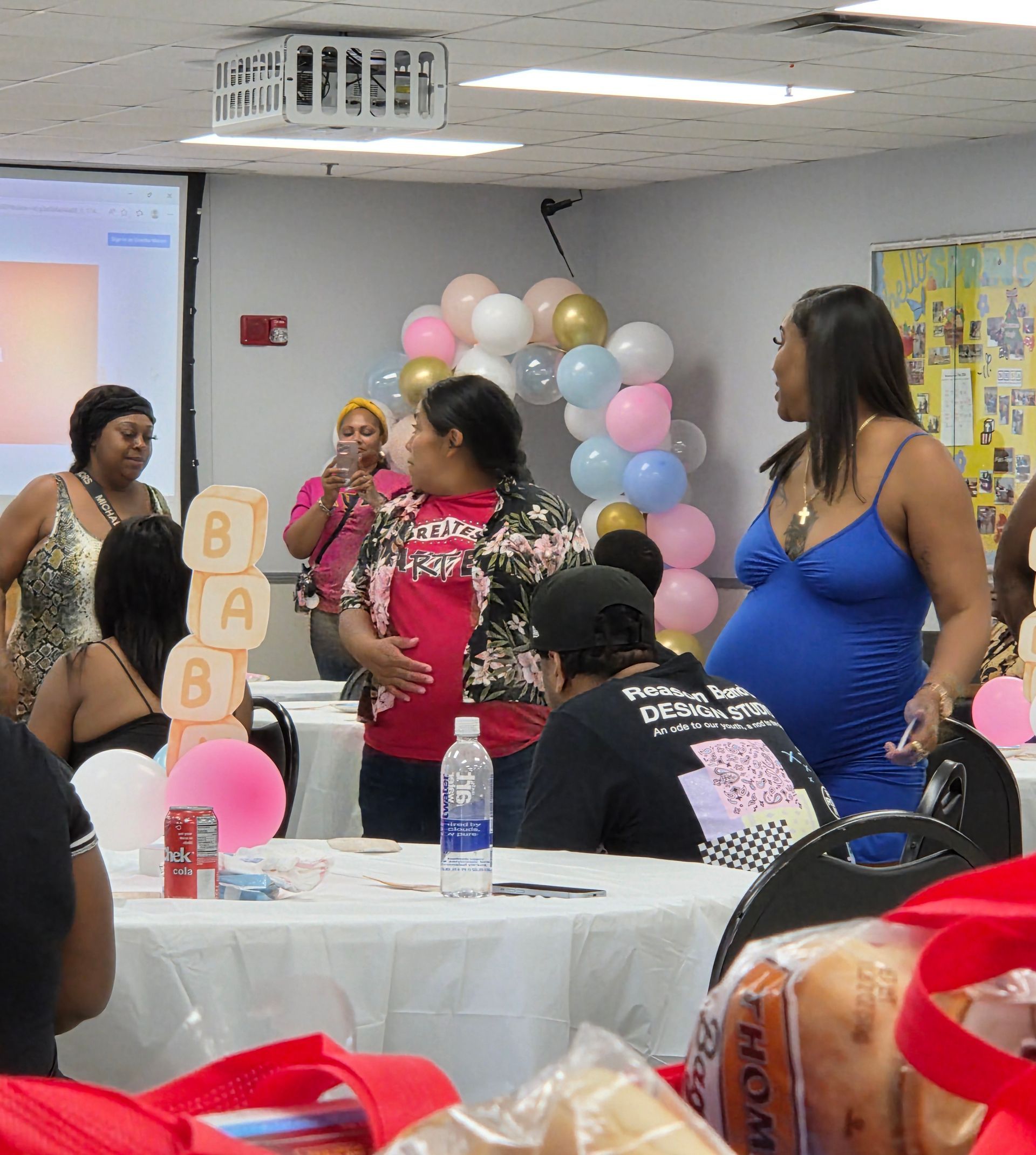 People gathered at a baby shower with pastel balloons, gifts, and a decorated table in a community room.