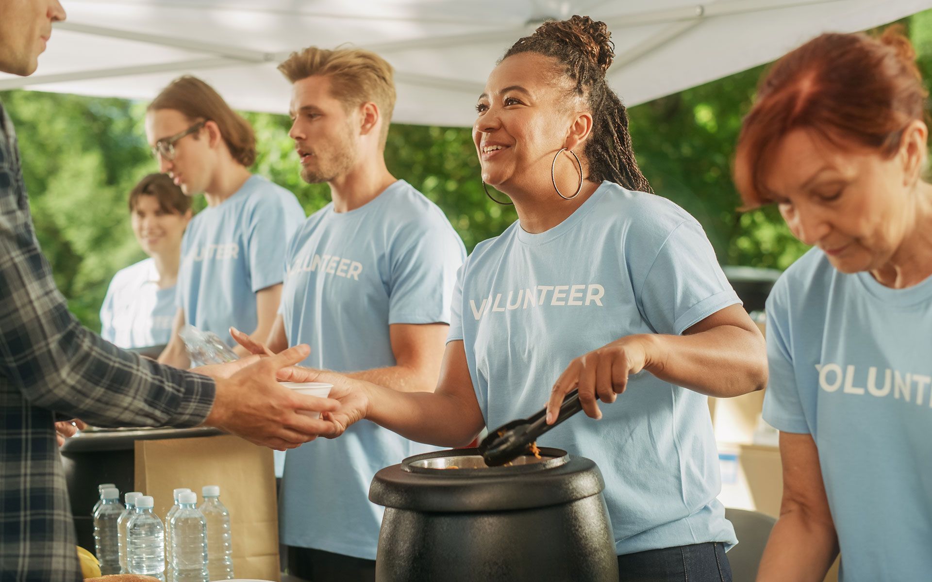 Volunteers in blue shirts serving food from a pot to a person outdoors.
