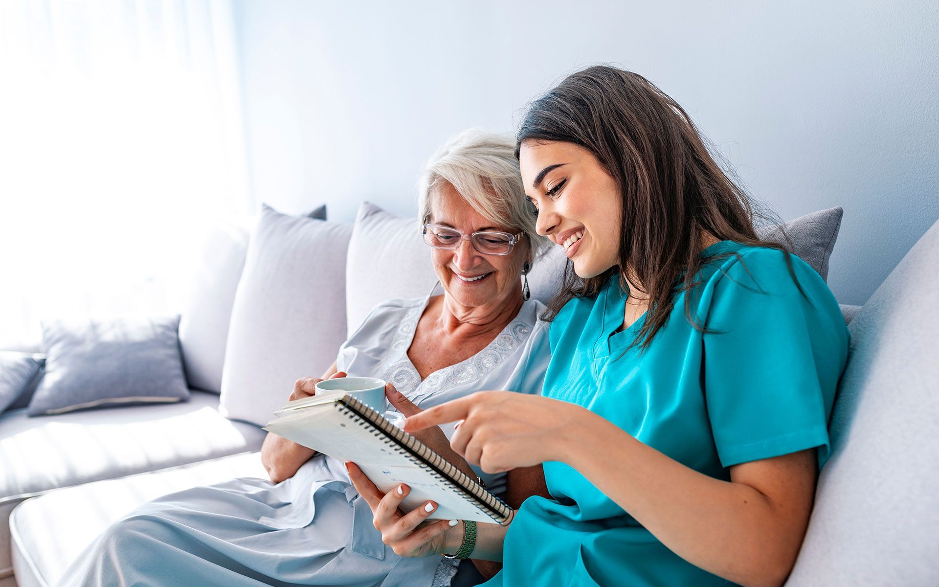 Woman in teal scrubs points at a tablet screen while seated next to an older adult woman on a couch.