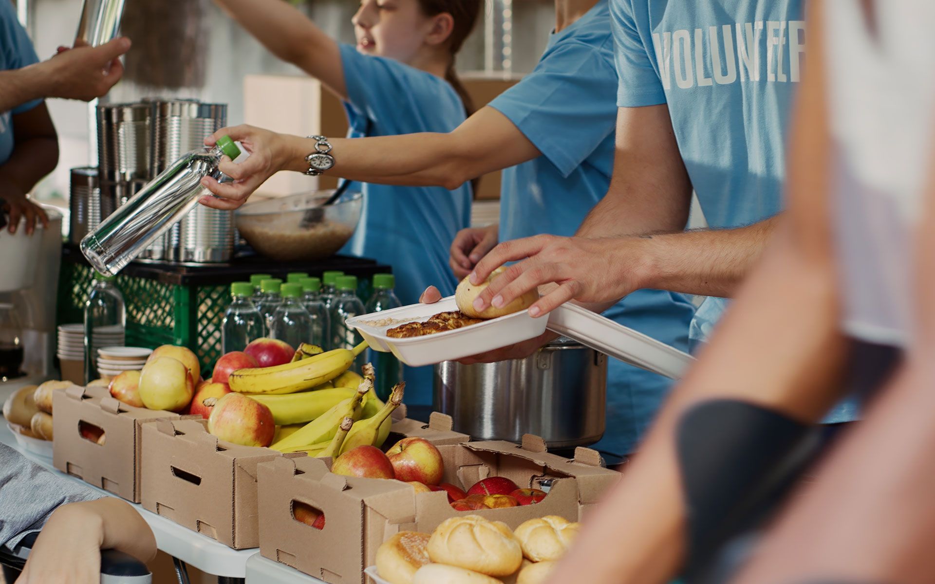 Volunteers serving food at a table with fruits, water bottles, and a container of soup.