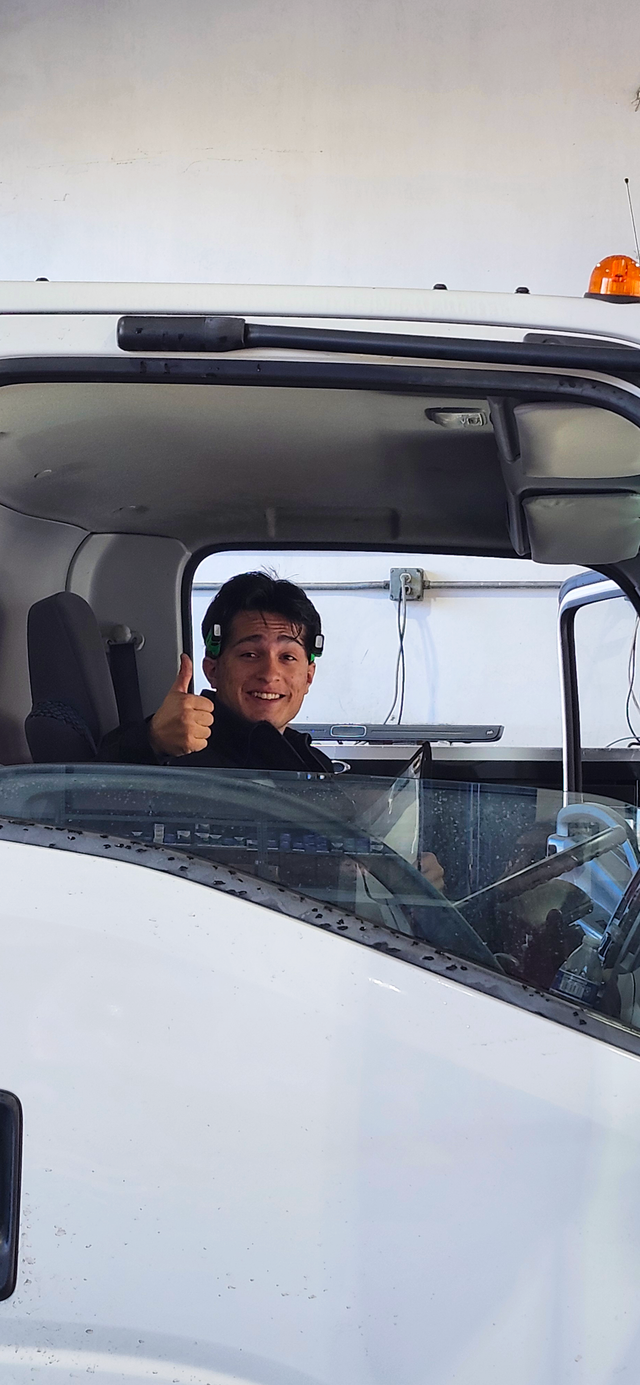 Person smiling and waving from inside a white truck cab with an open windshield area. | Redhawk Auto Service - Temecula