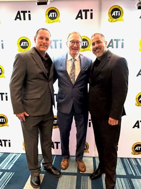 Three men in suits posing together in front of an ATI Excellence event backdrop.