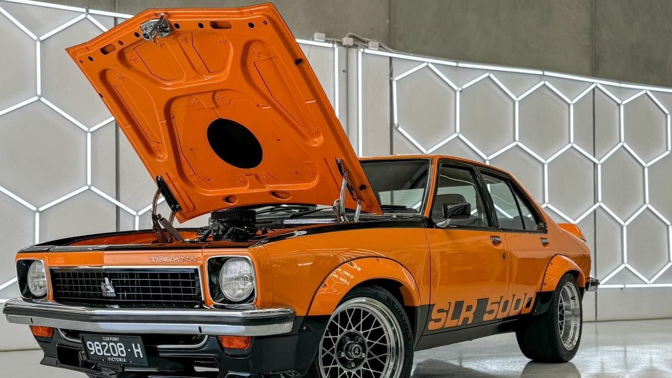 Orange Holden Torana SLR 5000 with hood open, parked in a studio with hexagon wall decor.