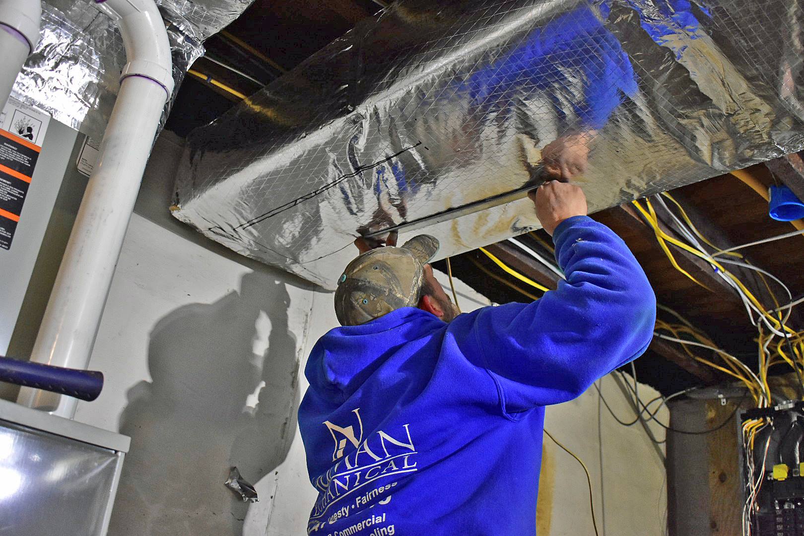 A man from Nauman Inc. in a blue hoodie is working on an air duct in a basement.