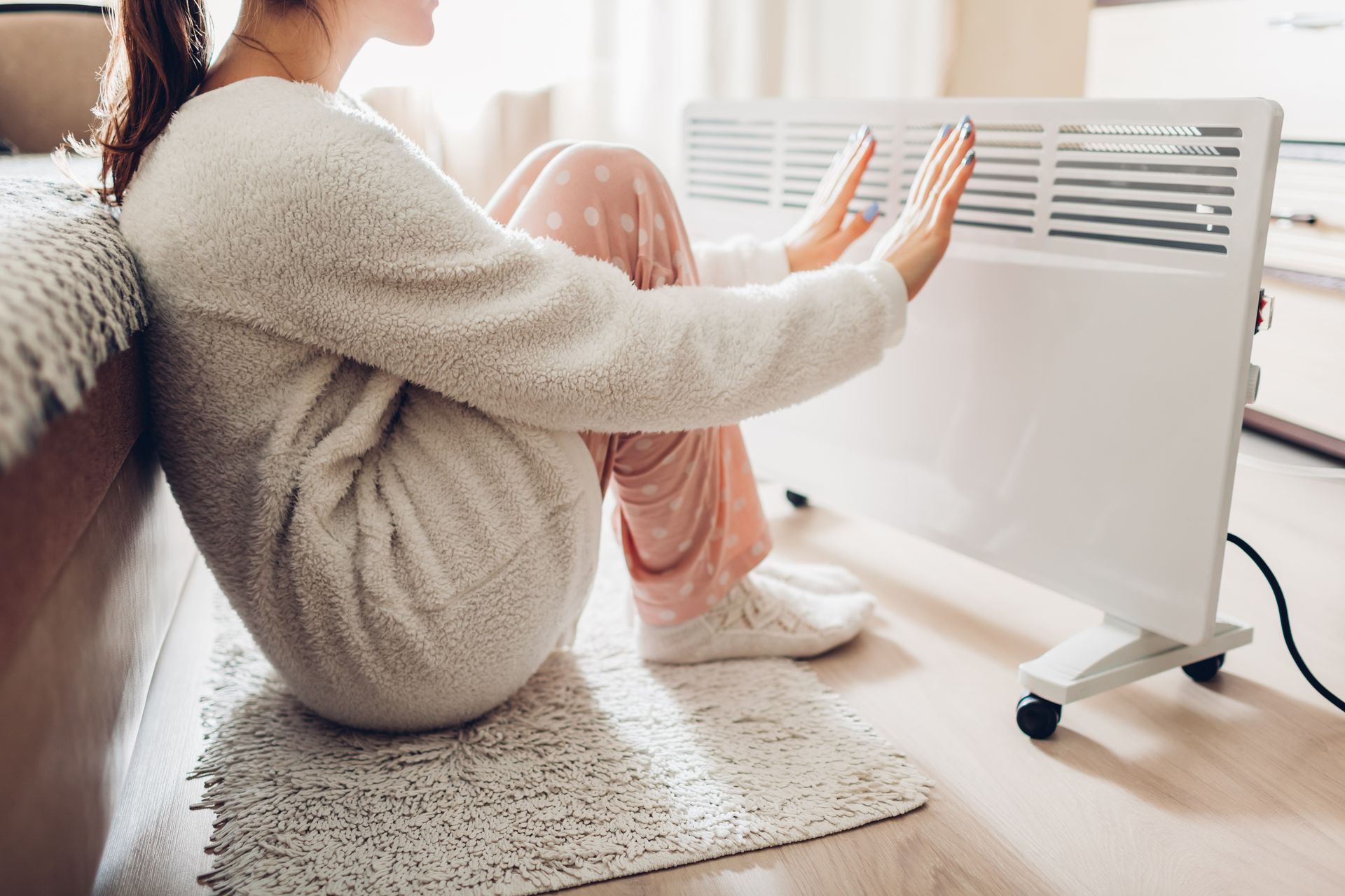 Woman in pajamas warming hands by a white electric heater on a rug.