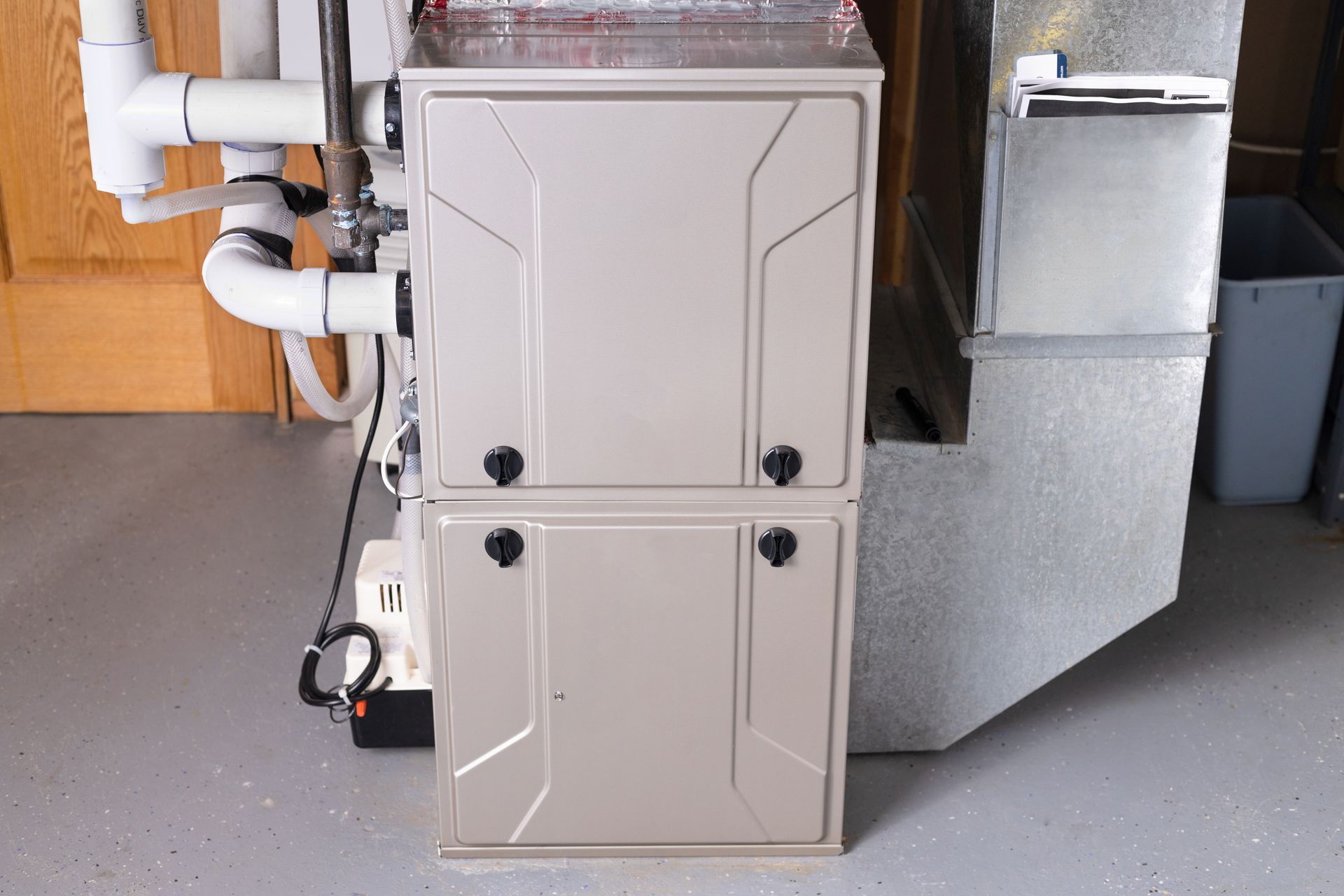 A beige furnace with two doors, next to a silver duct and white pipes, indoors.
