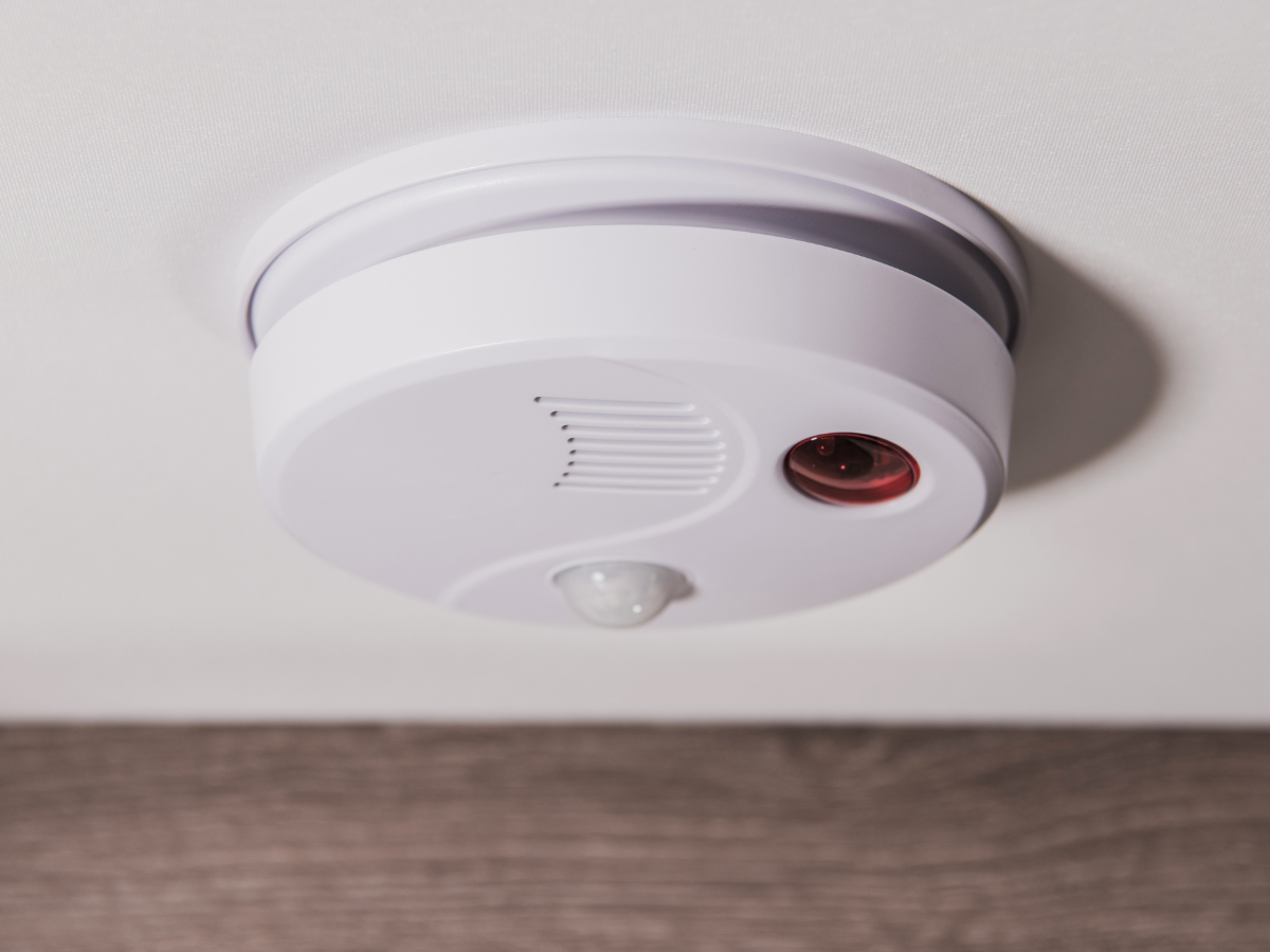 White smoke detector mounted on a white ceiling with a red indicator light.