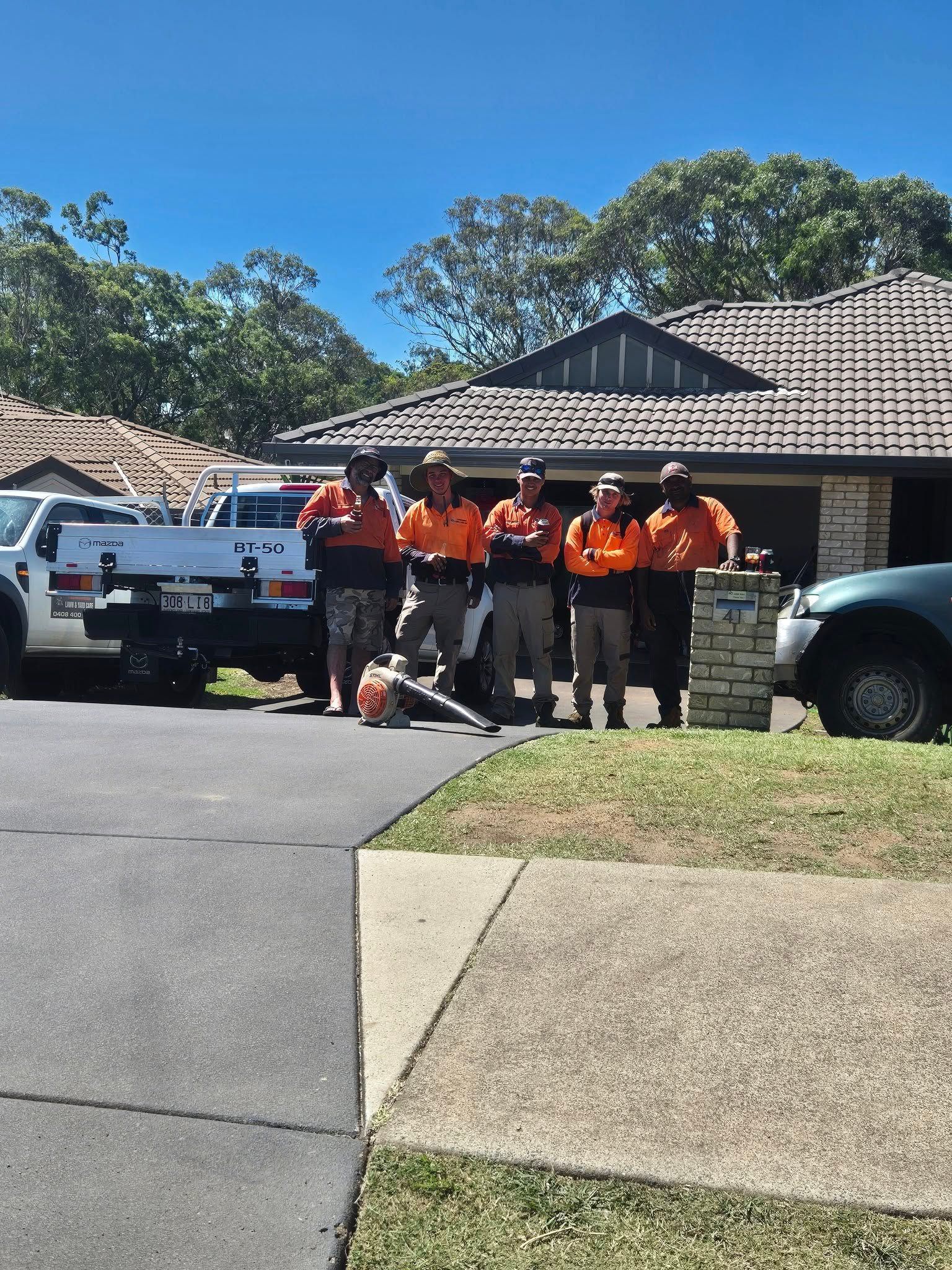 Workers in Orange Shirts Stand Outside a Suburban House Beside Utility Trucks — Toowoomba Lawn and Yard care In Glenvale, QLD