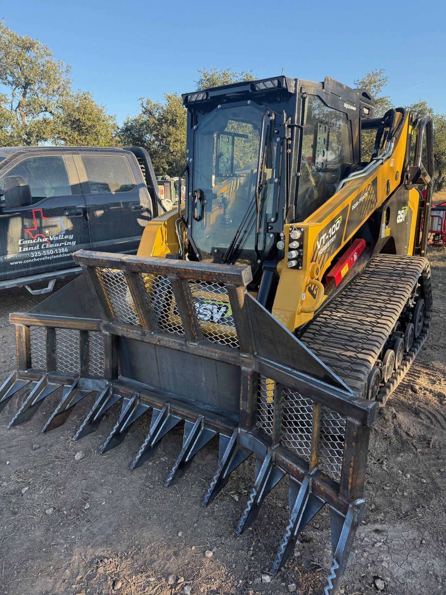 A yellow Caterpillar skid steer loader with a large, heavy-duty root rake attachment parked on a dirt lot.