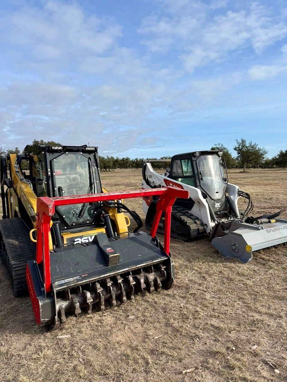 Two compact track loaders equipped with forestry mulcher attachments sit in an open, dry field under a blue sky.