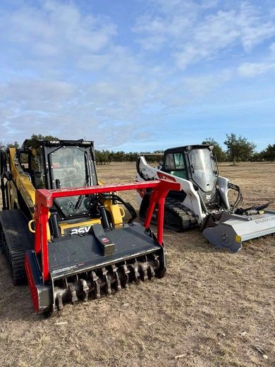Two compact track loaders equipped with forestry mulcher attachments sit in an open, dry field under a blue sky.