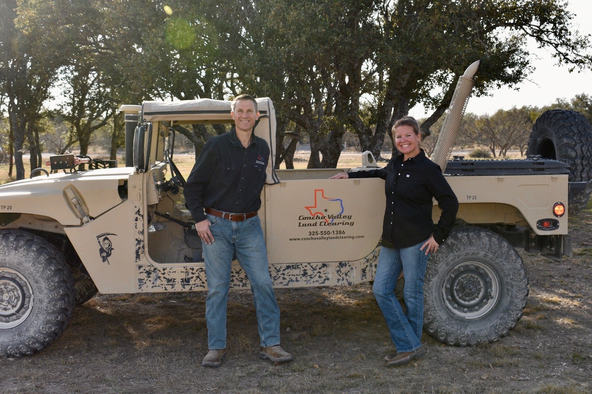 A man and woman stand smiling next to a tan, open-top military-style vehicle in a wooded outdoor setting.