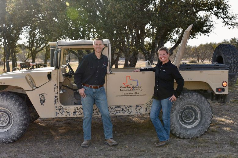 A man and woman stand smiling next to a tan, open-top military-style vehicle in a wooded outdoor setting.