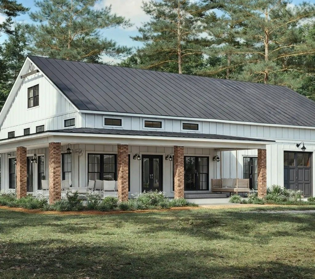 A white farmhouse with a dark metal roof and wraparound porch in a wooded yard.