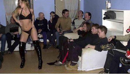 A woman in lingerie and tall black boots dances in front of a group of men seated on a couch and chairs.